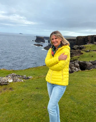 Susanne Eichhorn in gelber Jacke auf einer grünen Klippe am Meer